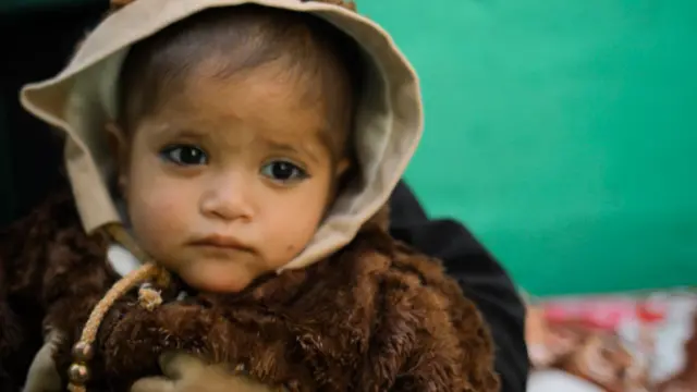 Una niña pequeña, vestida con un jersey marrón grueso y con una capucha beige en la cabeza, mira a la cámara