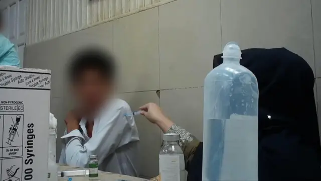 Una escena en un hospital. Una mujer, cuyo rostro no se ve, está poniendo una inyección a un paciente, cuyo rostro también está borroso. Le está poniendo la inyección sin guantes y clavándole la aguja a través de la manga de la camisa.