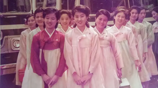 Una docena de jóvenes vestidas con trajes tradicionales coreanos posan delante de unos vehículos. Ko Yong-hui se encuentra en primera fila, a la izquierda. 
