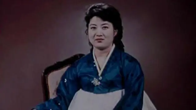 Ko Yong-hui, que debe de tener entre treinta y cuarenta años, lleva un vestido tradicional coreano azul y blanco y sonríe a la cámara