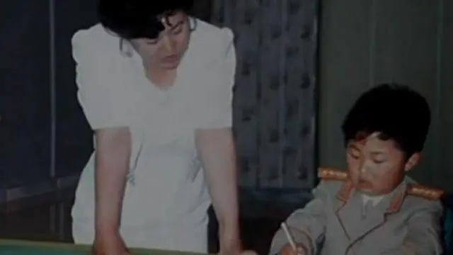 Ko Yong-hui, vestida de blanco, está de pie a la izquierda. Un joven Kim Jong-un, vestido con uniforme militar, está sentado y escribe con un bolígrafo. 